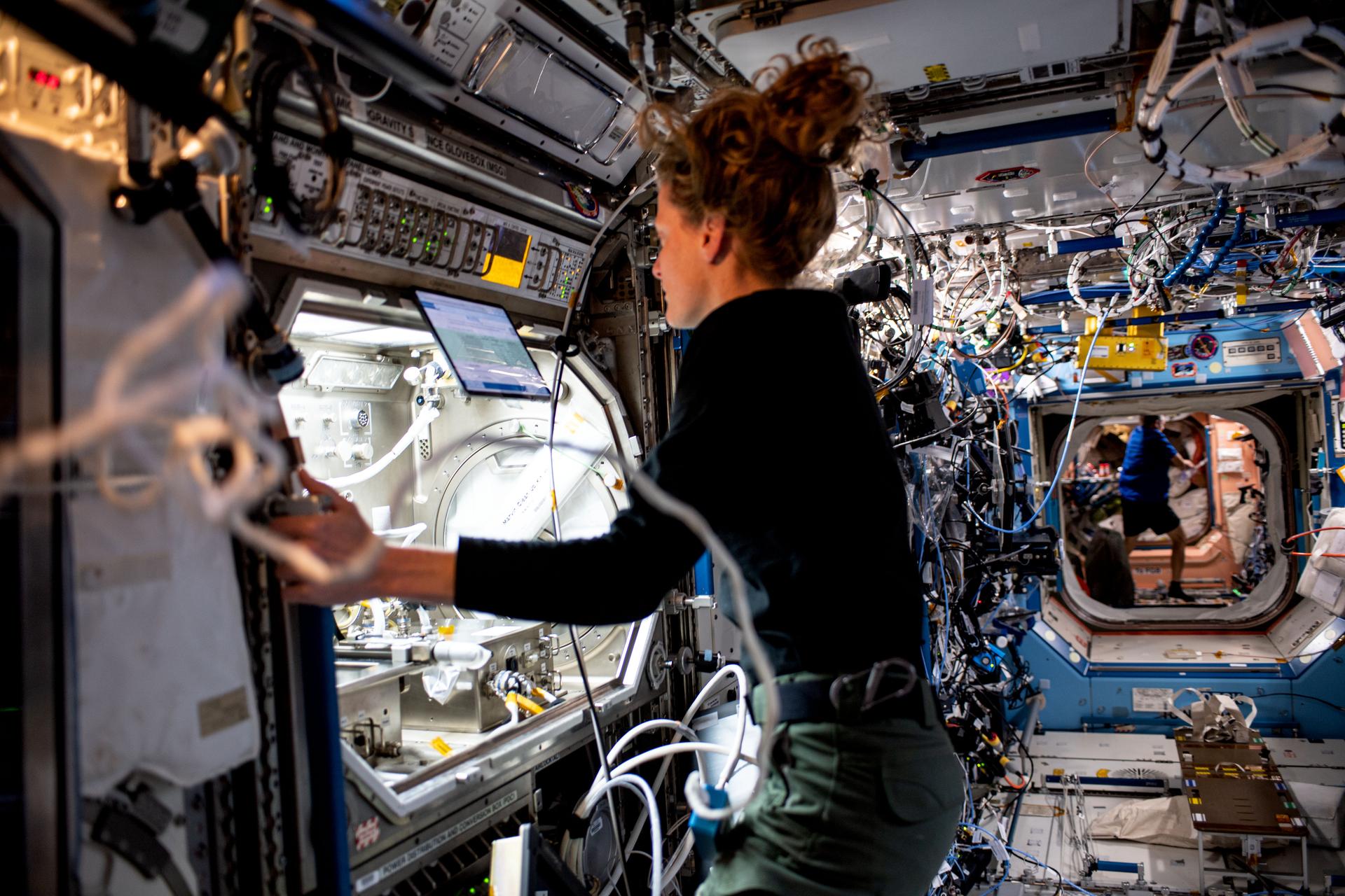 O’Hara is wearing a long-sleeved black shirt and green pants, with her hair in a ponytail. She is standing in front of the open front of a large, lighted module with two silver boxes inside it, each with multiple hoses and connectors. She is looking at a laptop screen at the top of the module and holding on to the edge of the hardware with her left hand.